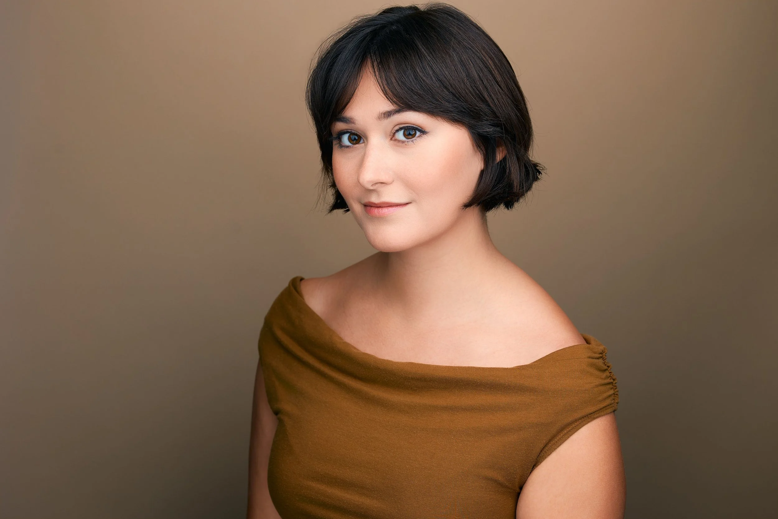 Studio headshot of actor Emily Michelle Walton wearing a golden-brown off-the-shoulder top and looking at the camera with a soft smile.