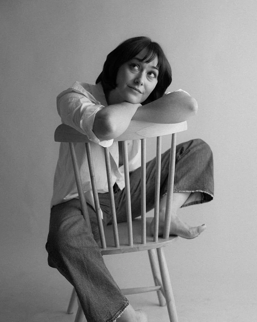 Black-and-white studio portrait of actor Emily Michelle Walton sitting backward on a wooden chair, looking thoughtfully upward.
