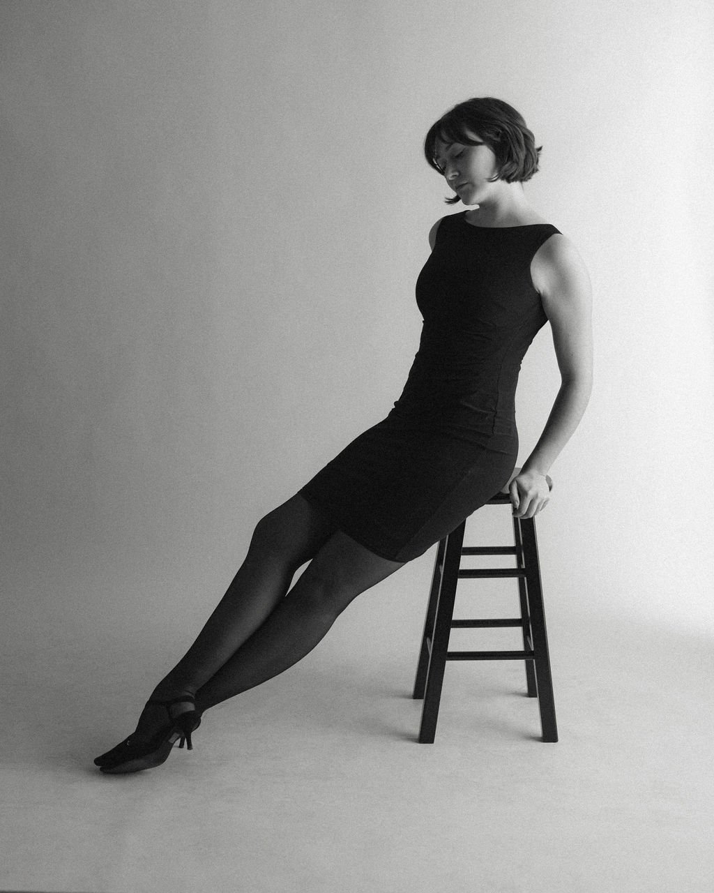 Studio portrait of actor Emily Michelle Walton sitting on a stool in a black dress, leaning to one side with her legs extended.