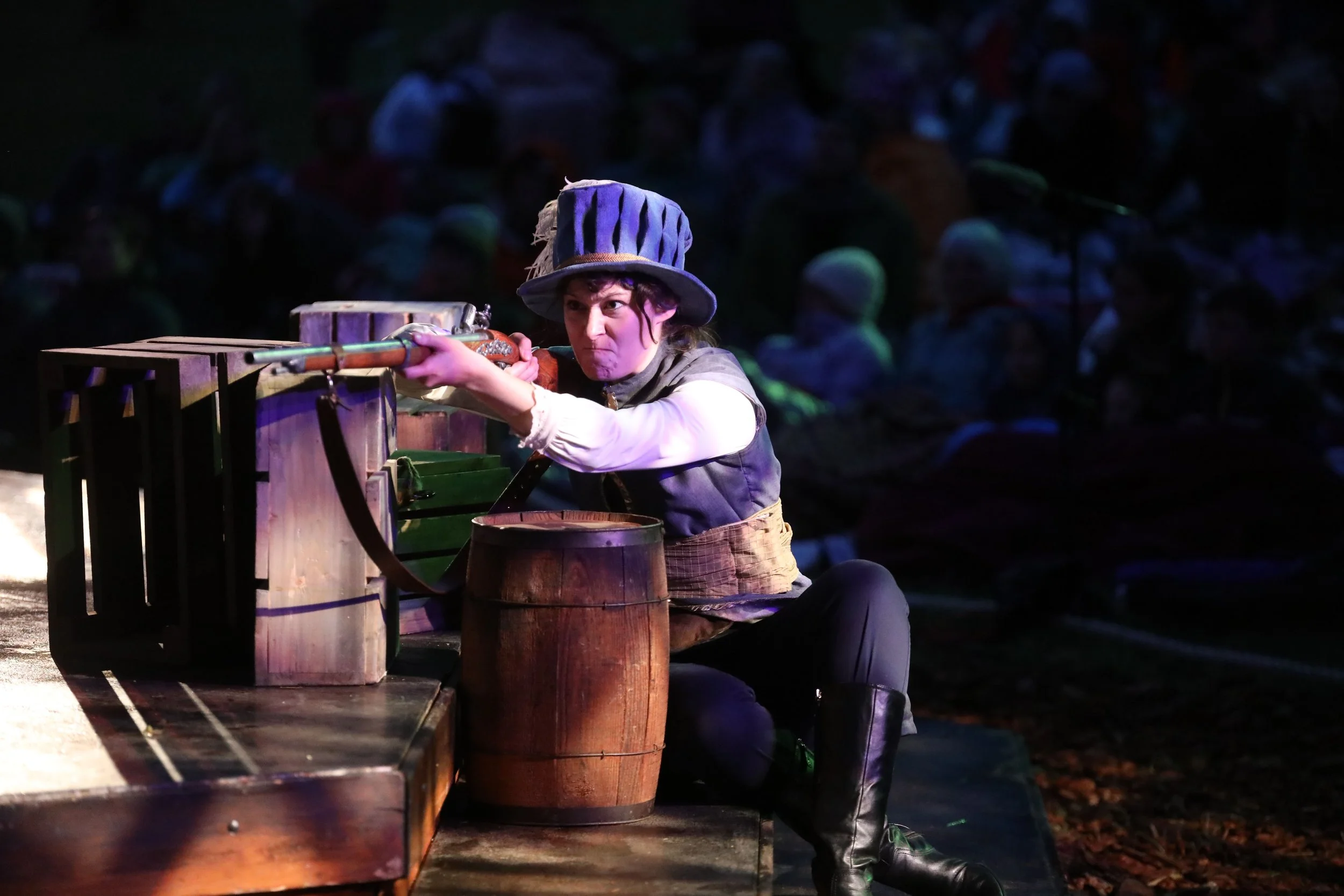 Emily Michelle Walton as a soldier aiming a rifle during the war scene in The Three Musketeers at Montana Shakespeare in the Parks, performing on an outdoor stage at dusk.