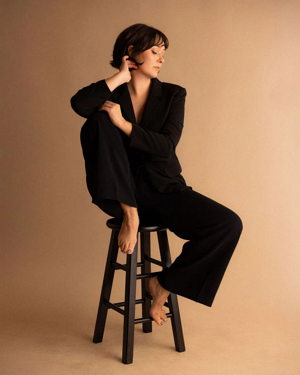 Studio portrait of actor Emily Michelle Walton sitting on a black stool in a black suit, one leg crossed over the other with her eyes closed.