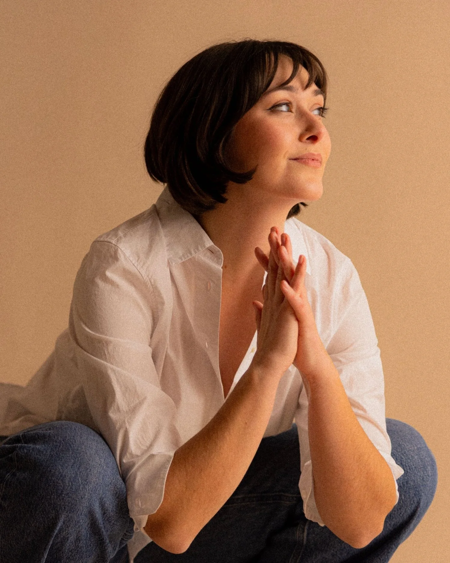 Studio portrait of actor Emily Michelle Walton sitting cross-legged on the floor, wearing a white shirt and blue jeans and looking thoughtfully to the side