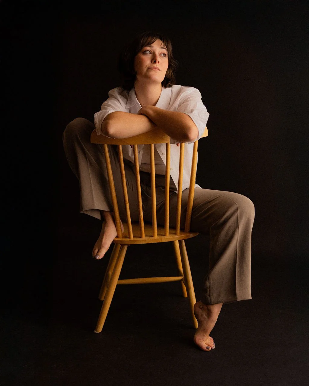 Studio portrait of actor Emily Michelle Walton sitting backward on a wooden chair with her arms resting on the backrest, looking thoughtfully to the side.