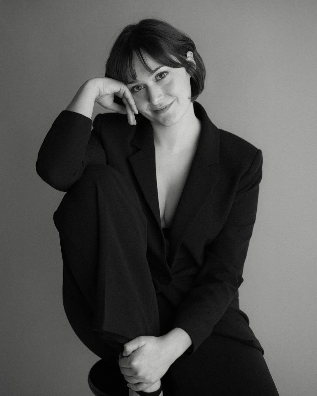 Black-and-white studio portrait of actor Emily Michelle Walton wearing a blazer, seated with one knee raised and smiling softly at the camera.