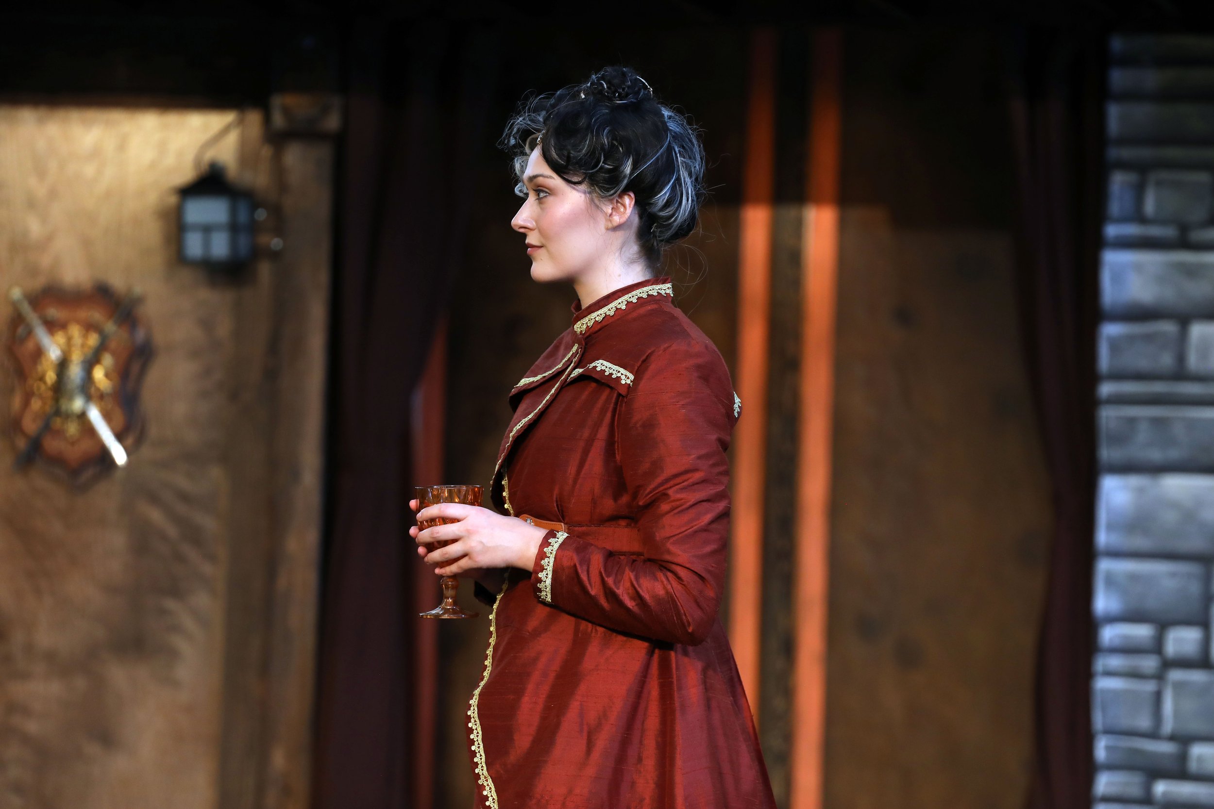 Emily Michelle Walton as Gertrude in Hamlet at Montana Shakespeare in the Parks, standing in profile holding a glass, wearing a red gown with gold embroidery.