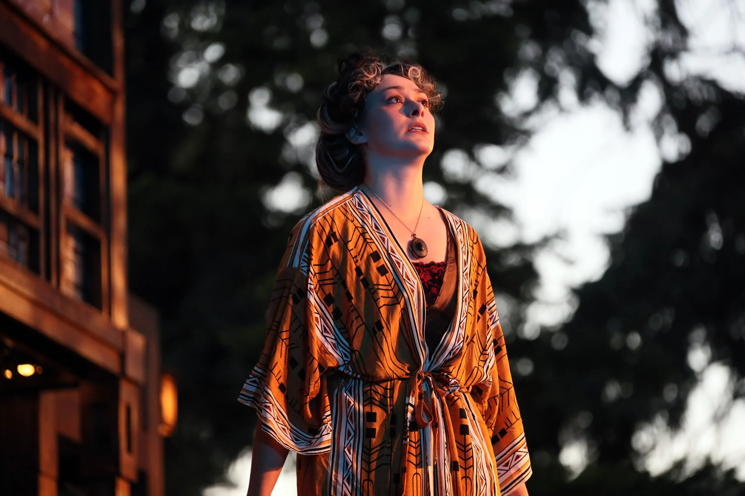 Emily Michelle Walton as Gertrude in Hamlet at Montana Shakespeare in the Parks, wearing a patterned robe and standing outdoors at sunset.