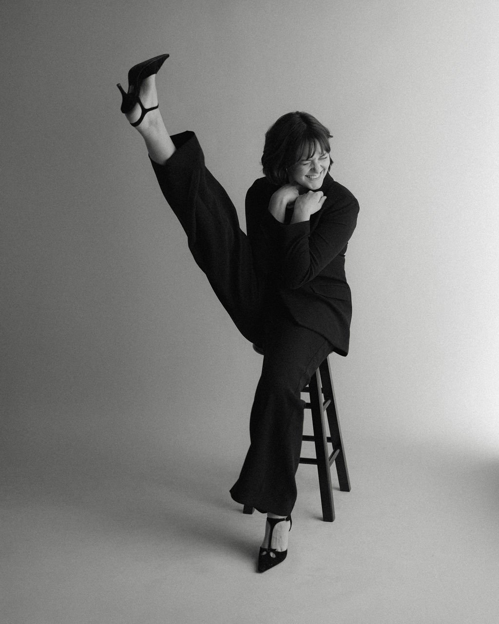 Black-and-white studio portrait of actor Emily Michelle Walton sitting on a stool with one leg raised in a playful high kick, smiling with her eyes closed.