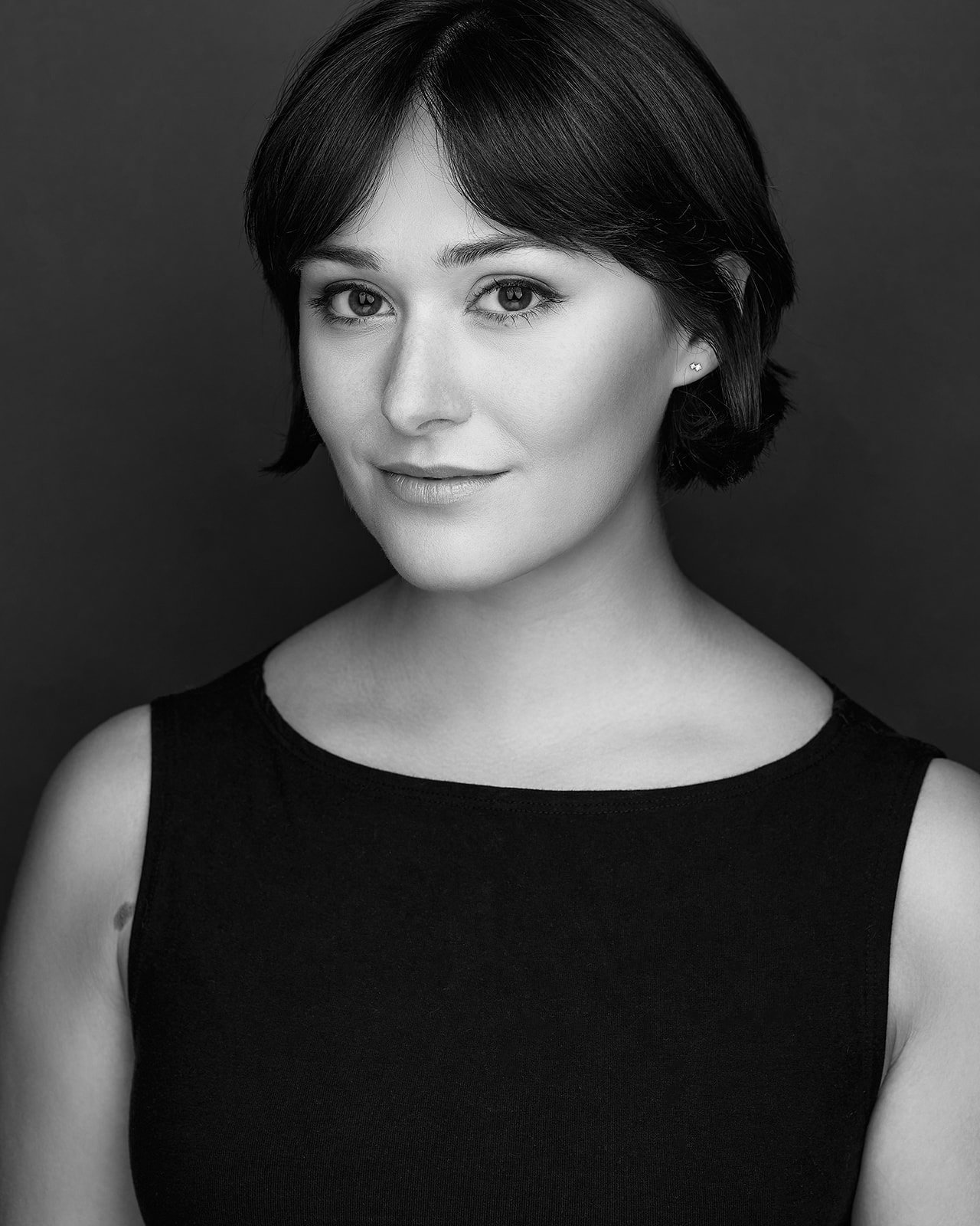 Black-and-white studio headshot of actor Emily Michelle Walton in a sleeveless black top against a dark background, smiling subtly.