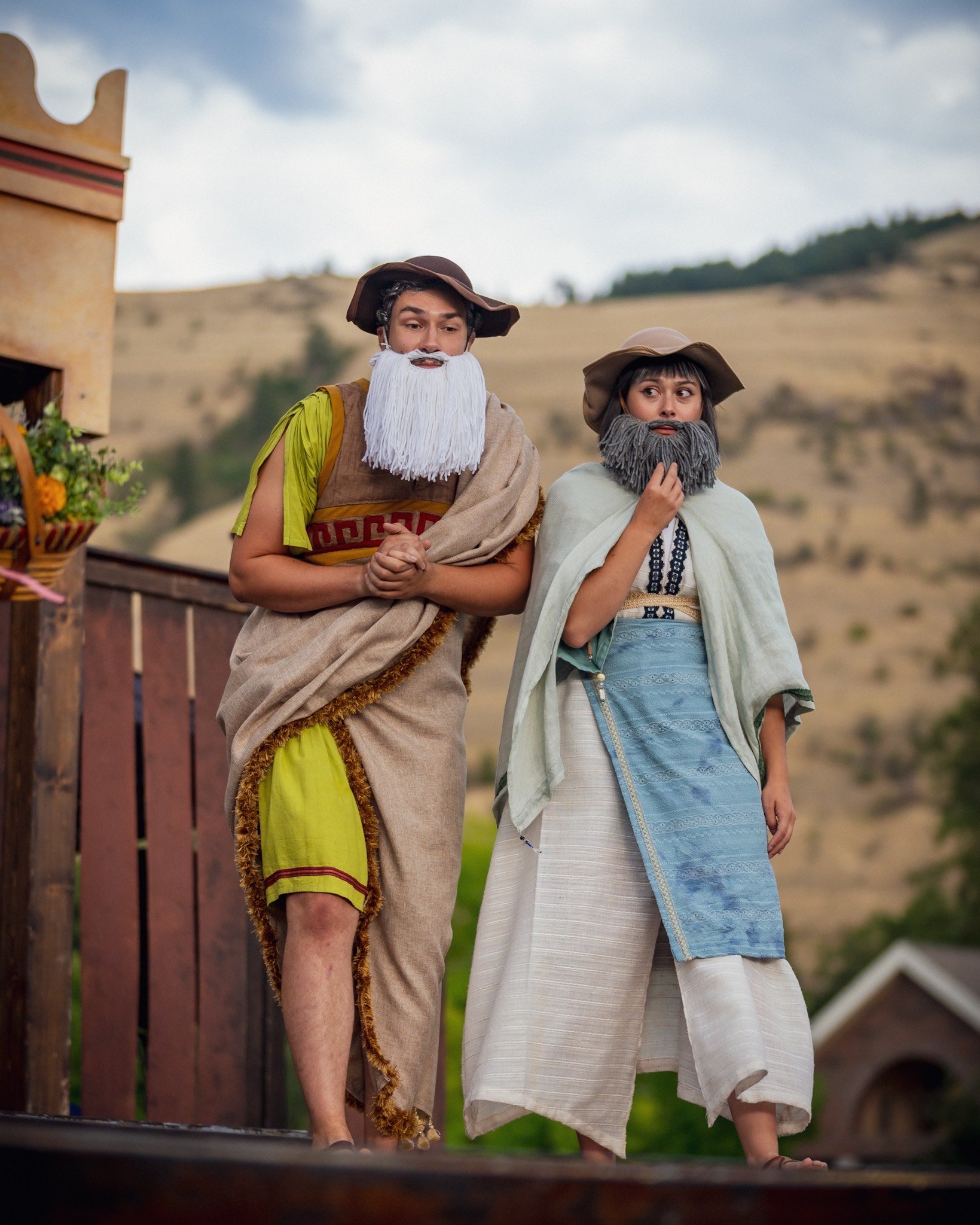 Emily Michelle Walton as the disguised Camillo and Calvin Adams as the disguised Polixenes in The Winter's Tale at Montana Shakespeare in the Parks, standing outdoors in Mycenaean-inspired costumes and false beards.