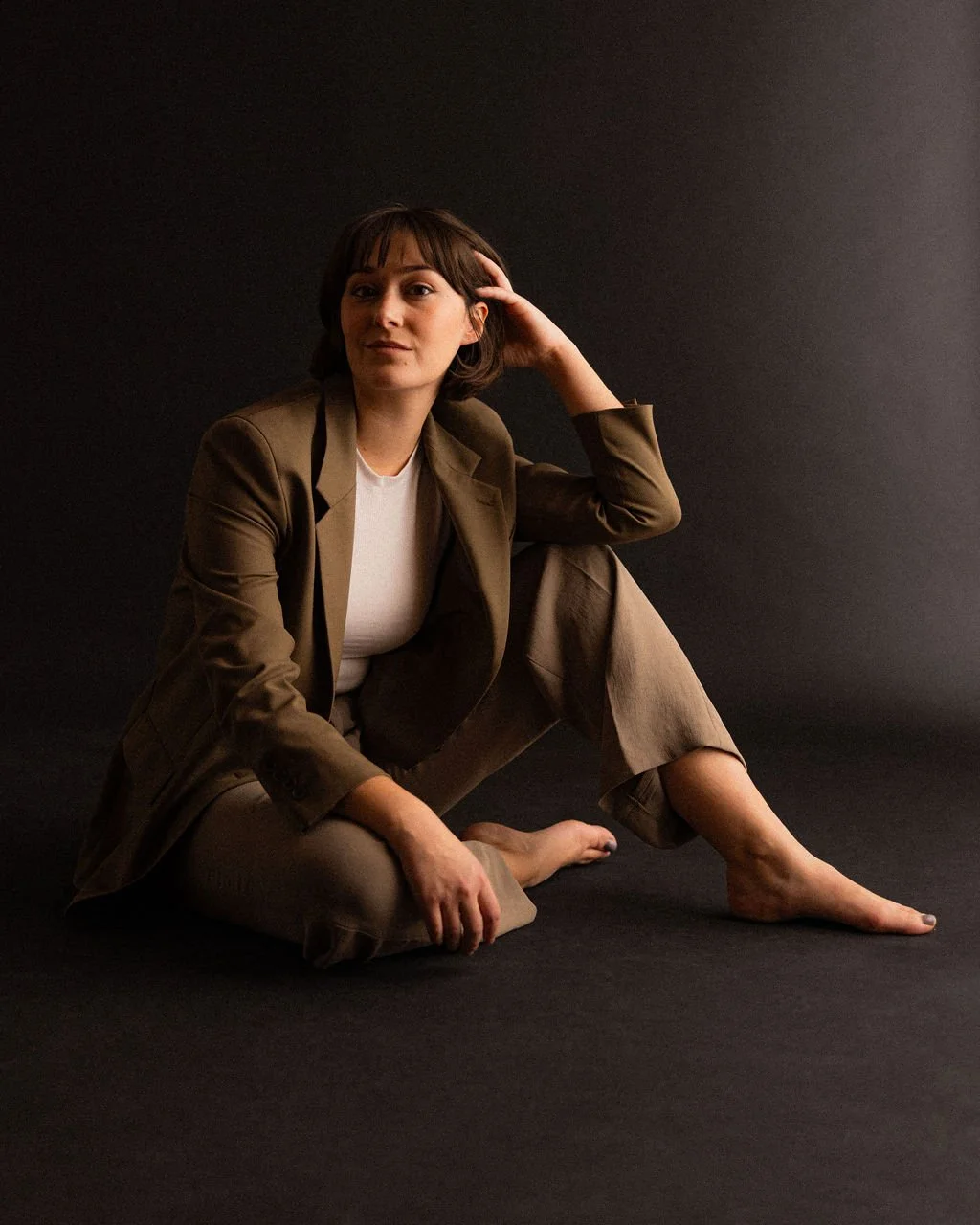 Studio portrait of actor Emily Michelle Walton wearing a brown blazer and trousers, seated on the floor against a dark background and looking at the camera.