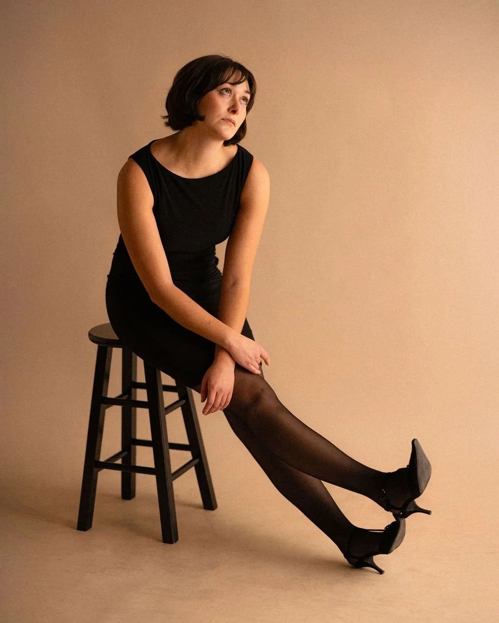 Studio portrait of actor Emily Michelle Walton seated on a stool with her legs extended, leaning forward slightly and looking upward with a playful, bored expression.