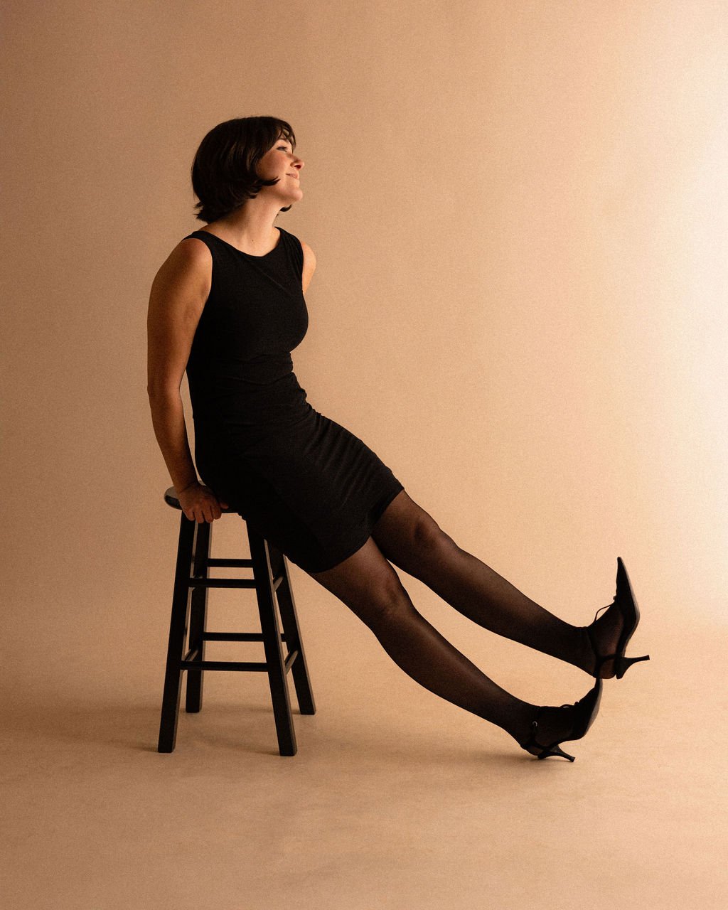 Studio portrait of actor Emily Michelle Walton seated on a stool with her legs playfully extended and stacked at the ankles, leaning forward and smiling while looking slightly upward.