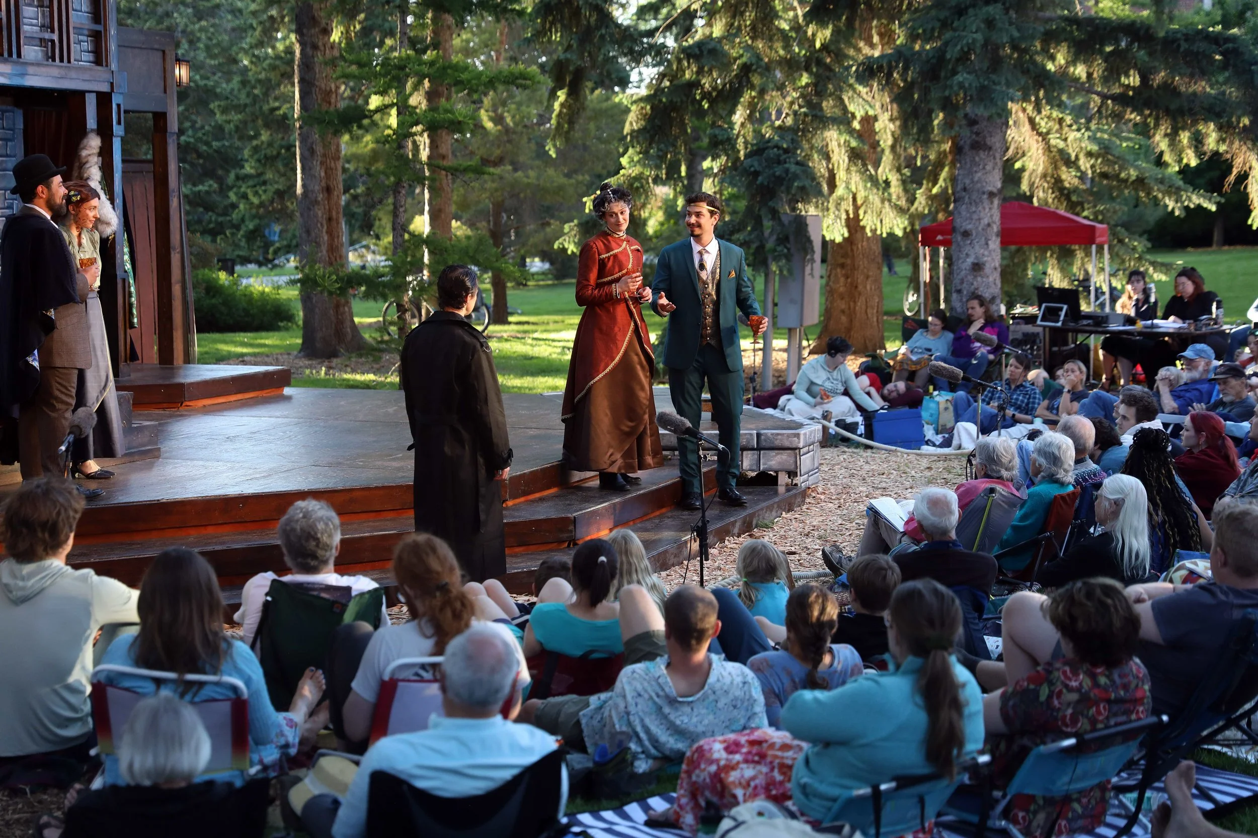 Wide production shot of Hamlet at Montana Shakespeare in the Parks, with Gertrude, Claudius, and Hamlet performing on an outdoor stage before an audience at MSU's Duck Pond in Bozeman.
