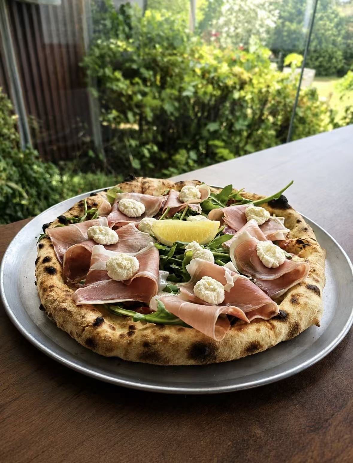 Pizza topped with prosciutto, dollops of cheese, arugula, and a lemon wedge on a metal tray, with greenery outside in the background.