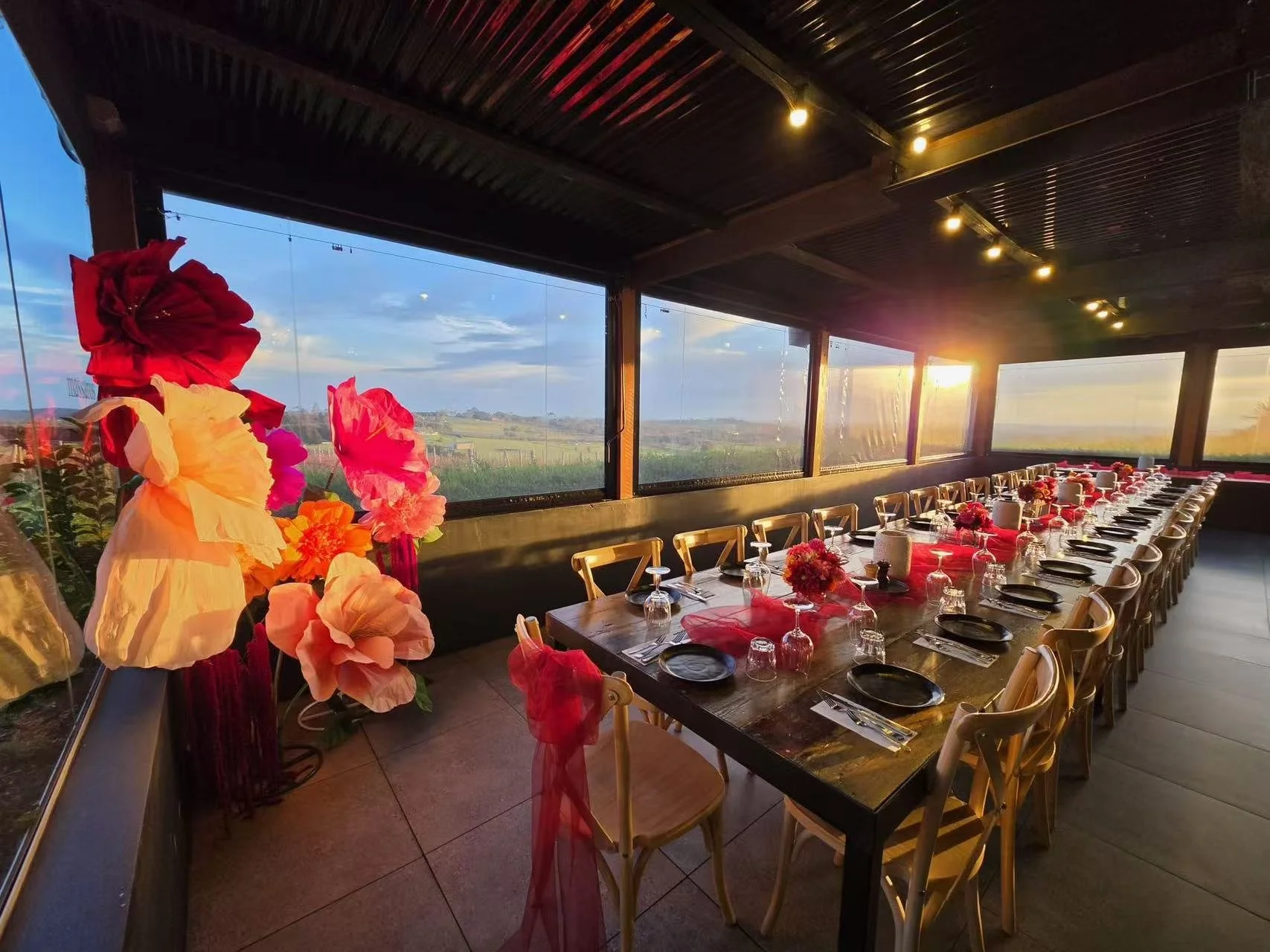 A long dining table with place settings and floral centerpieces, decorated for a special occasion, with large windows showing a sunset landscape outside, and flower arrangements along the windowsill.