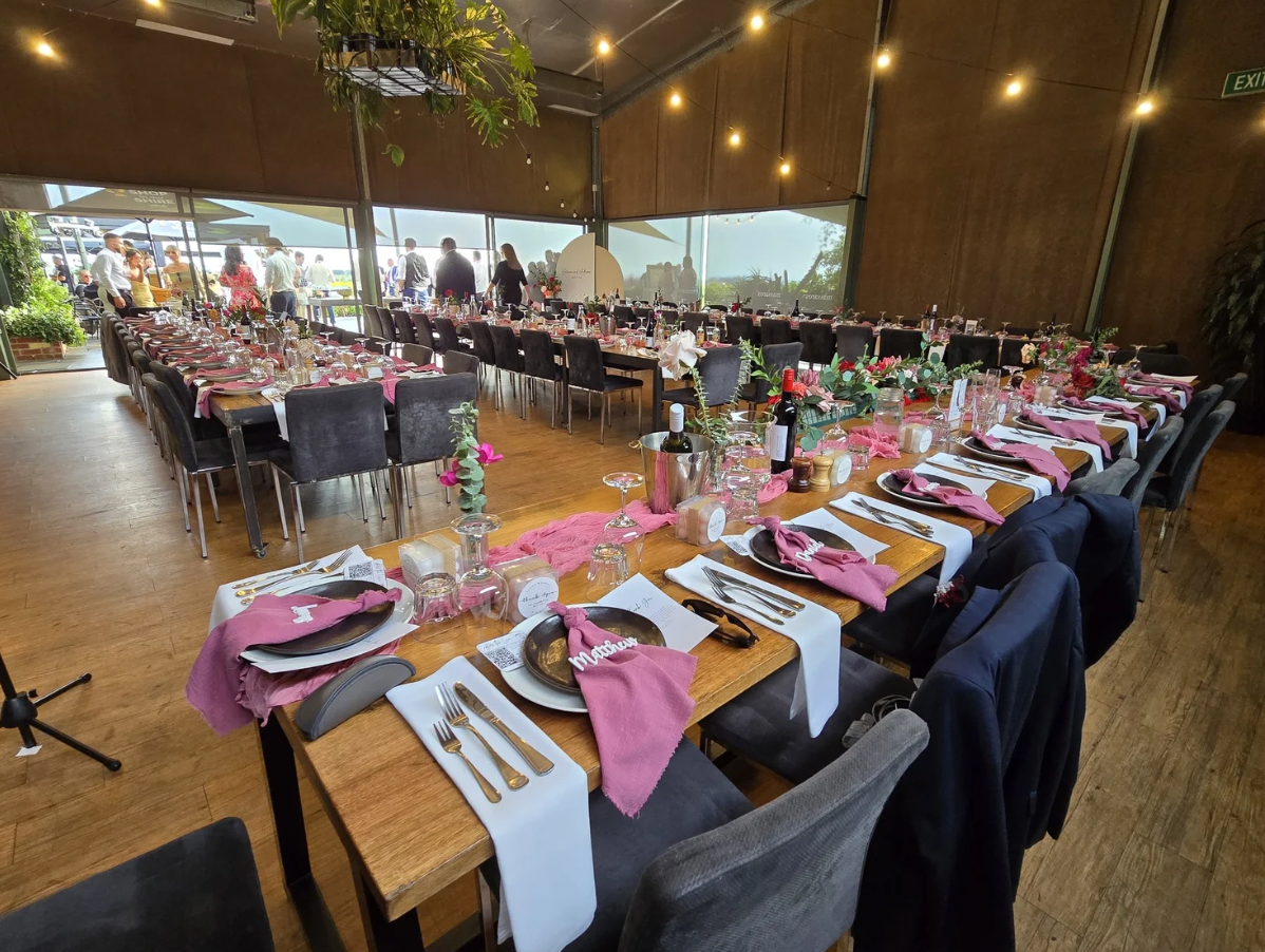 Wedding reception setup with long tables decorated with pink napkins, flowers, wine bottles, and place settings, in a spacious room with large windows and hanging string lights.