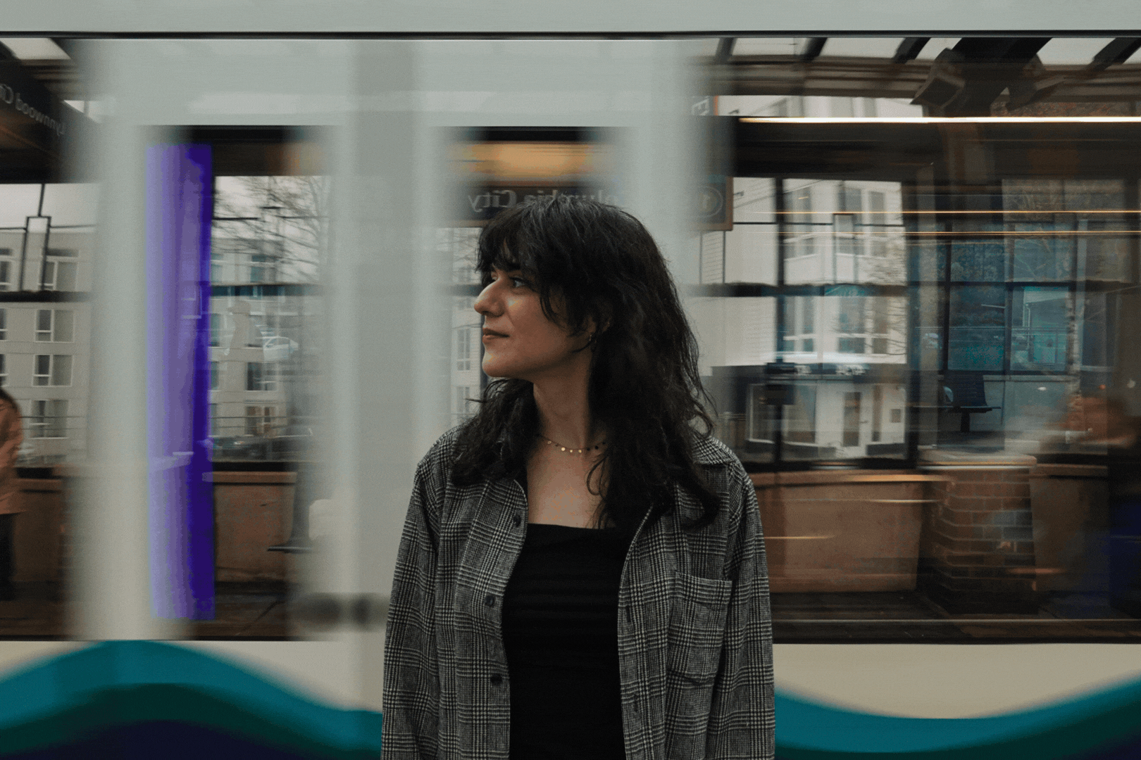 A woman with curly dark hair standing on a train platform, looking to her side. Behind her, a light rail train passes her in a blur.