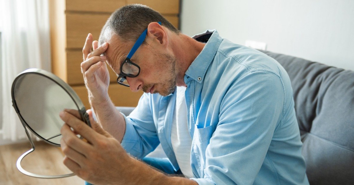 A middle-aged, balding man inspecting the loss of his hair in a small mirror he is holding in his hand.