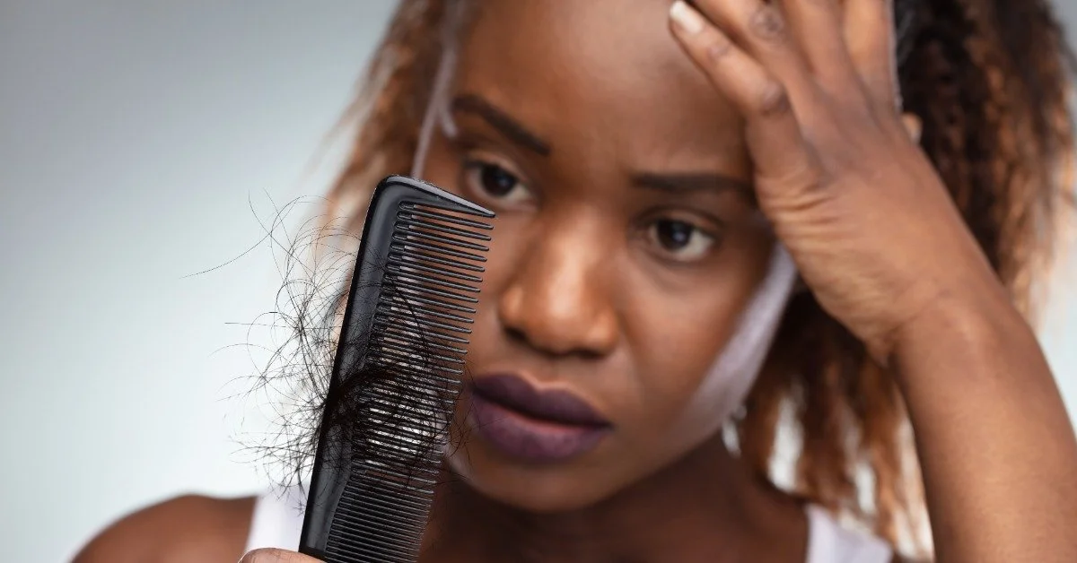 A woman looking worriedly at a comb in her hand covered in her hair. She is holding her head with her other hand.