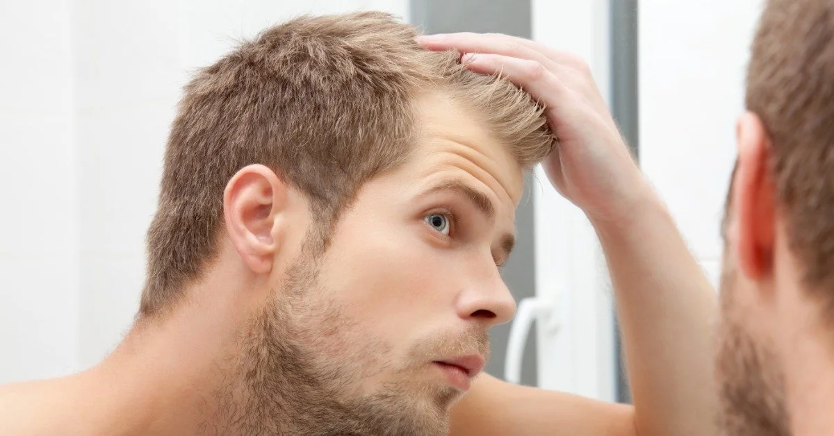 A young man looking a mirror as he pulls his hair back to inspect if his hairline is receding at all.