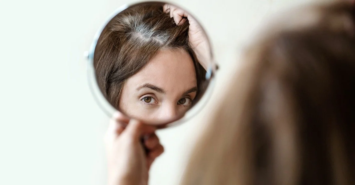 A woman holding a small hand mirror. She is using the mirror to inspect the part of her hair that is thinning.