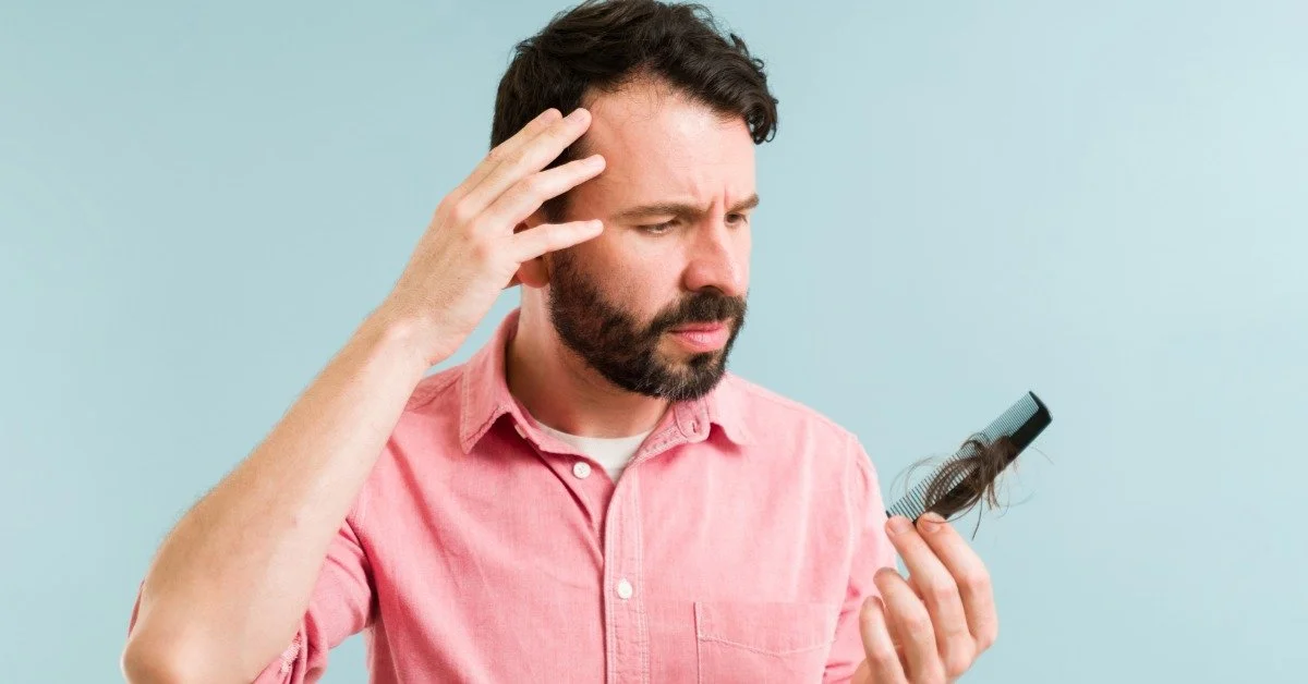 A man looking worriedly at a comb in his hand. There is a clump of hair that has fallen out of his hairline in the comb.