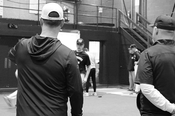 Baseball players practicing in an indoor training facility with netting and a staircase in the background.