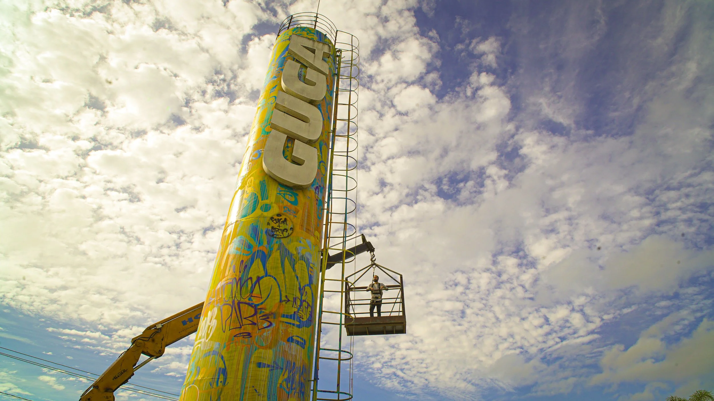 Pilar de vidro com letras grandes que dizem 'CUIABÁ', em uma altura elevada, com uma pessoa em uma plataforma de elevador suspenso ao lado, sob um céu com nuvens.