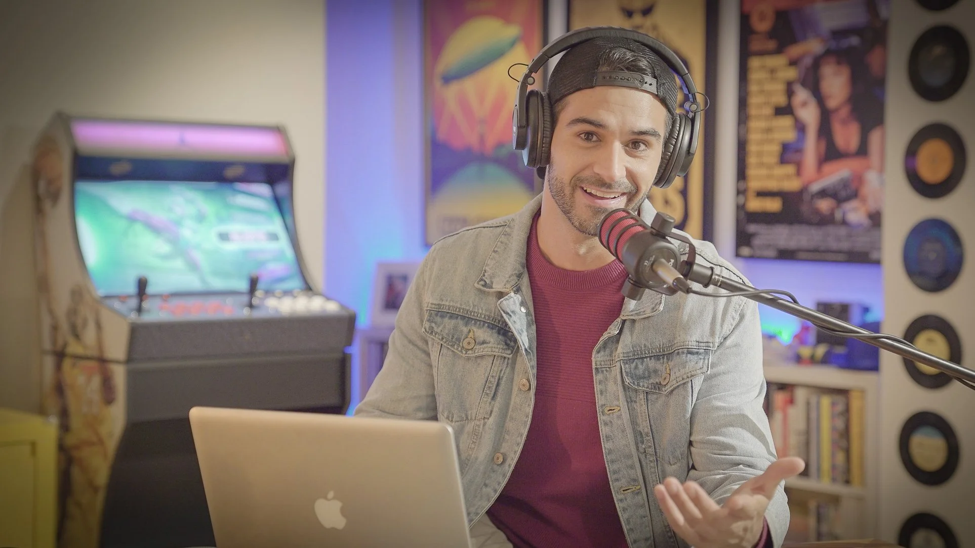 Homem sorridente usando fone de ouvido com microfone, sentado na frente de um laptop Apple, na frente de uma parede decorada com pôsteres e discos de vinil, em um estúdio caseiro de gravação ou podcast.