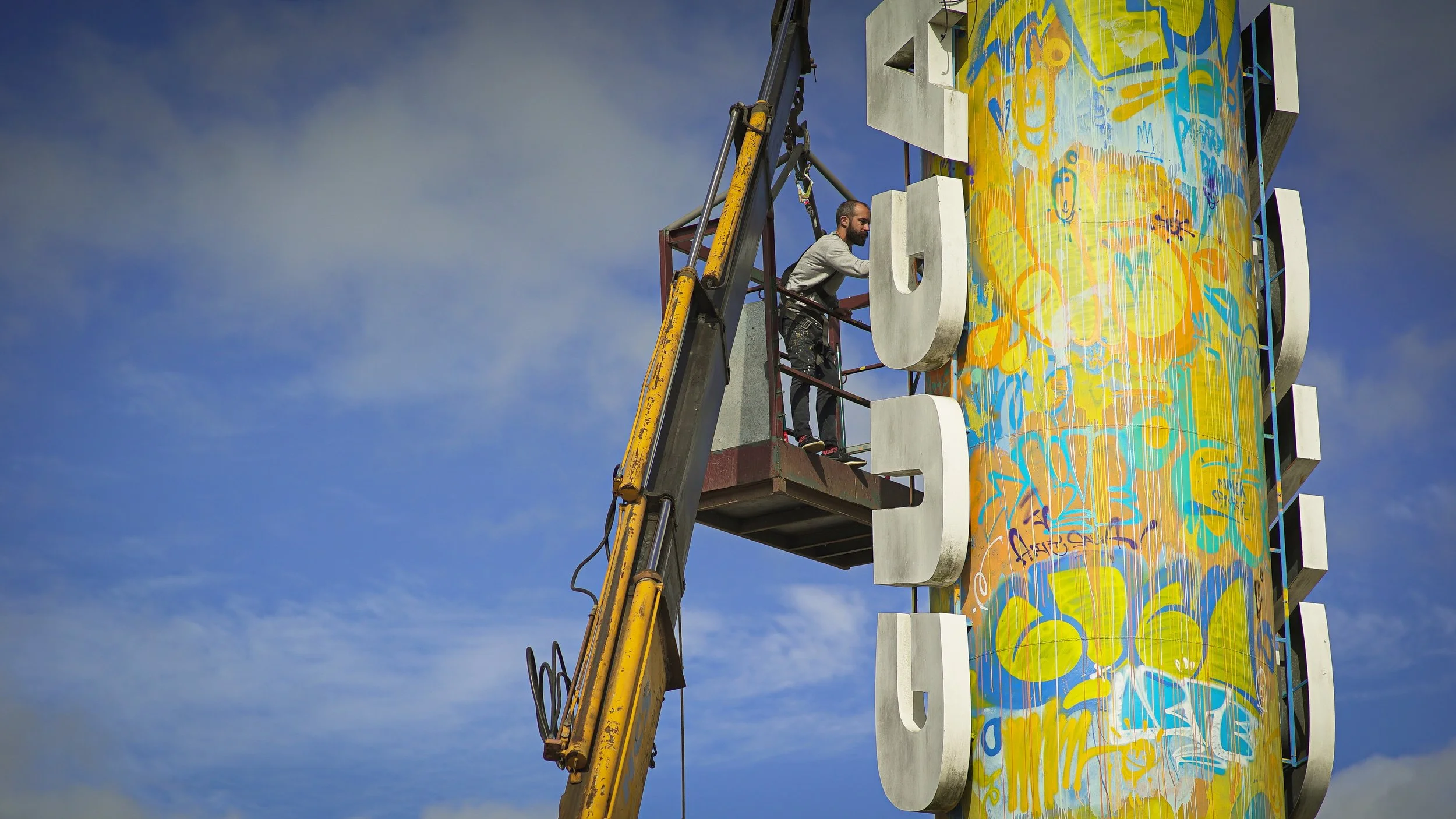 Pessoa em uma plataforma elevatória ao lado de um grande painel de arte urbana com letras em relevo.