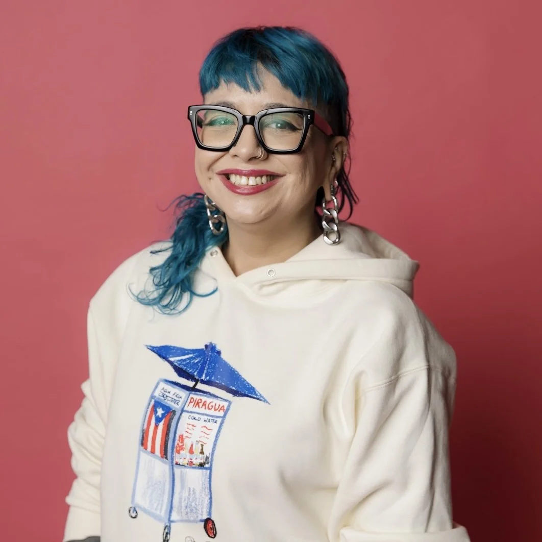 A person with tattoos and a smartwatch posing with a hand under their chin, smiling, wearing a black top.