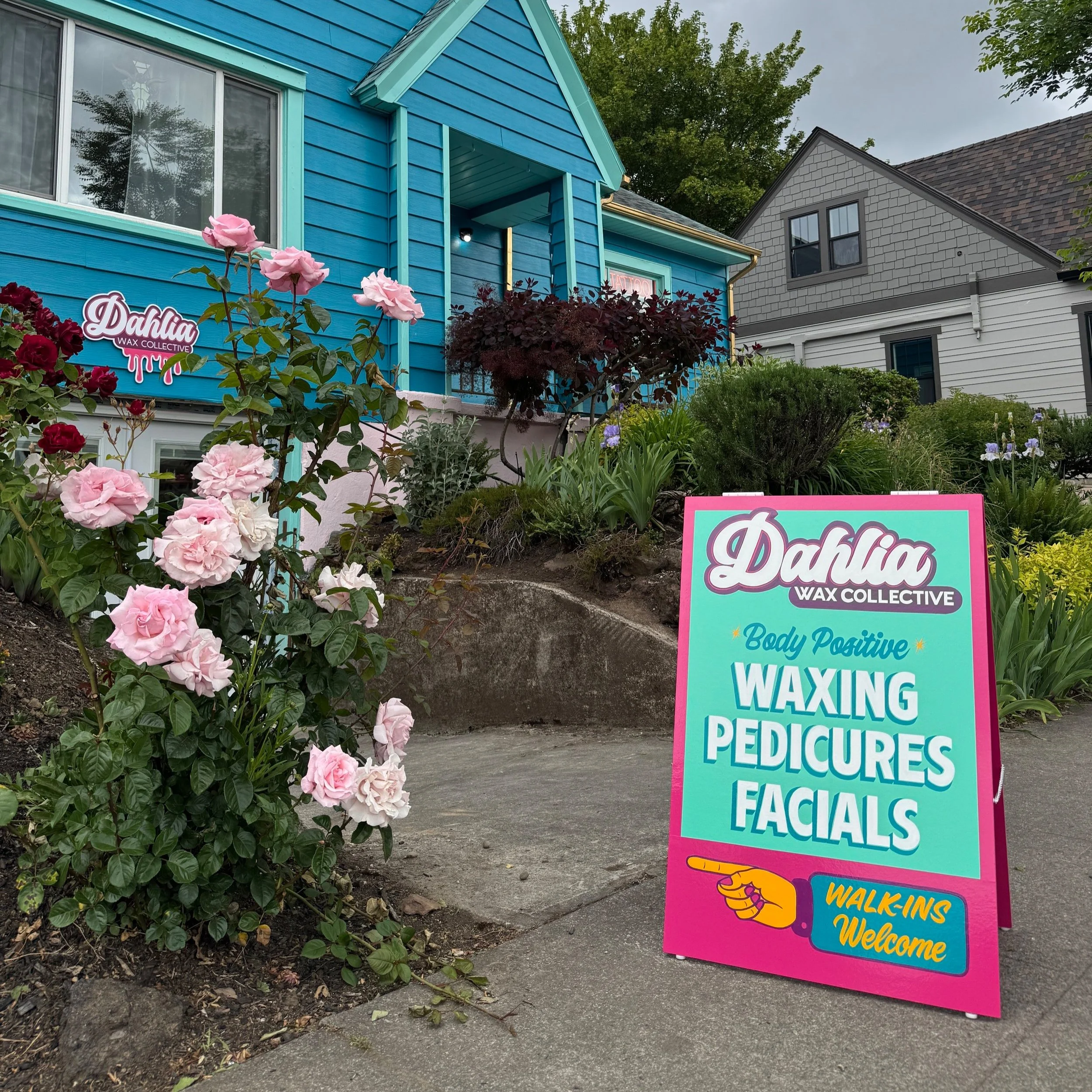 Sign outside a beauty salon for waxing services, with pink and teal colors, featuring the salon's name 'Dahlia Wax Collective' and mentioning body positive waxing, pedicures, and facials; house with blue exterior and garden with pink roses in the background.