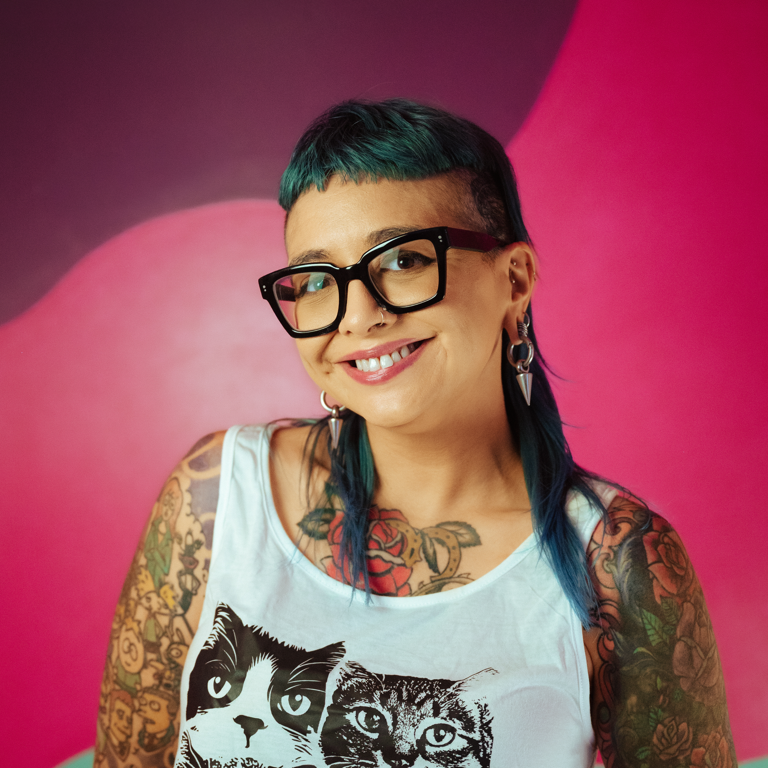 A person with tattoos and a smartwatch posing with a hand under their chin, smiling, wearing a black top.