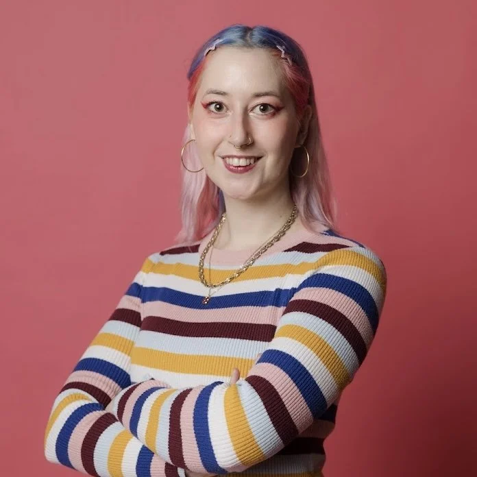 Portrait of a smiling woman with blonde hair, wearing a black t-shirt, with tattoos on her arms, standing against a neutral background.