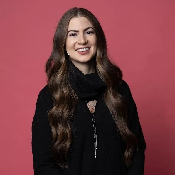 Smiling woman with long brown hair, wearing a black top, necklace, and showing tattoos on her arms.