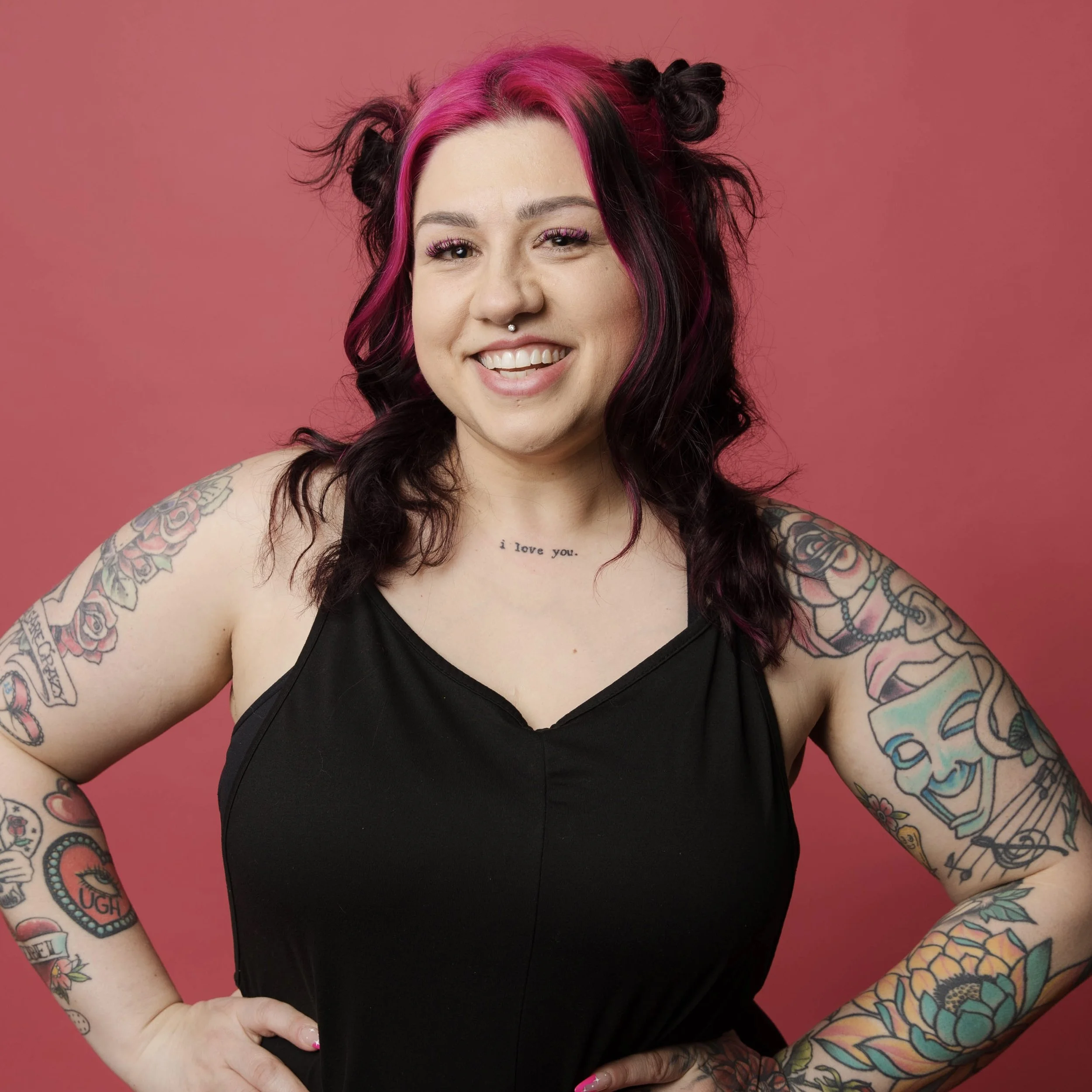 Person with pink hair and facial piercings, wearing a sheer black top, featuring tattoos on the arm and chest, smiling at the camera against a light background.