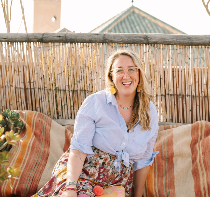 A smiling woman with glasses, yellow earrings, and long wavy hair sitting on a colorful outdoor sofa with striped cushions, in front of a bamboo fence and a house with a tiled roof.