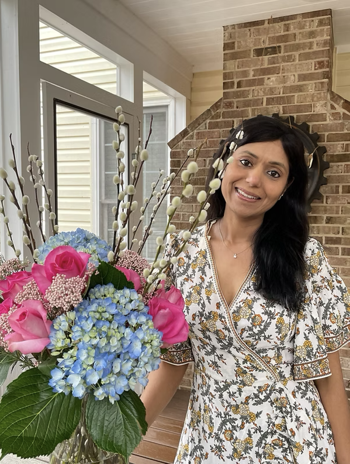 A woman with long dark hair wearing a floral dress standing indoors next to a bouquet of pink roses, blue hydrangeas, and pussy willow branches, with a brick fireplace and a glass door in the background.