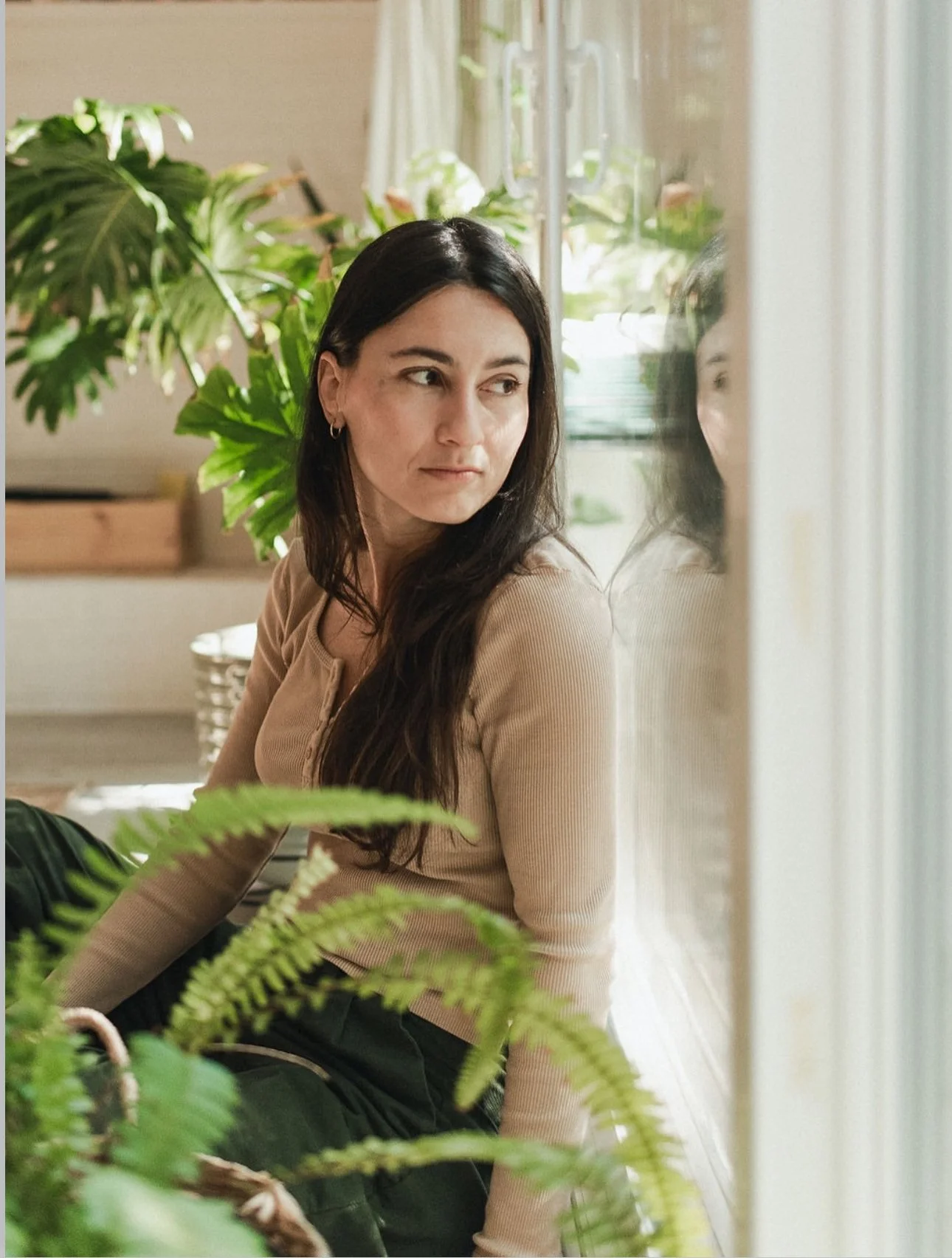 A woman with long dark hair sitting near a window surrounded by green plants, looking to her right.