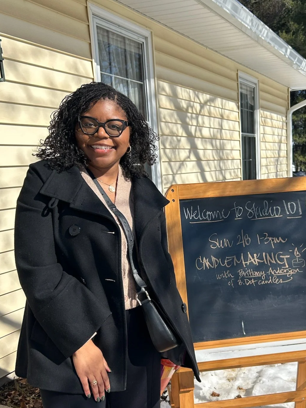 A woman with curly black hair, glasses, and earrings stands outdoors in front of a yellow house with white trim. She is smiling and wearing a black coat. Next to her is a blackboard sign with white writing, announcing a candle making event on Sunday, February 28th from 1 to 3 pm, hosted by Brittany Anderson of B.D. Candles.