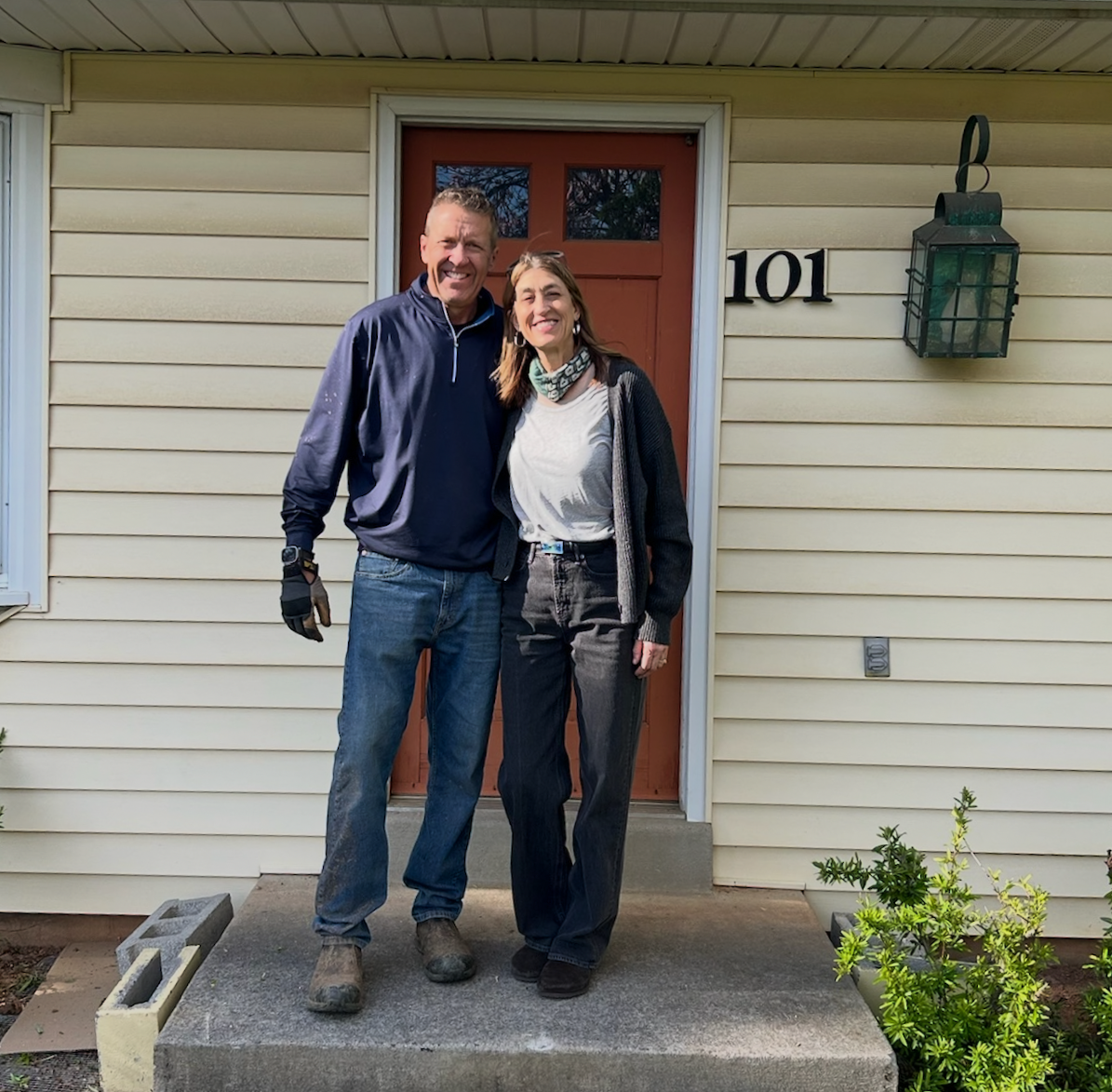 A man and woman standing on front porch steps smiling at camera in front of house with beige siding and red door, house number 101 displayed on wall, plants in planters, and outdoor lantern at side of door.