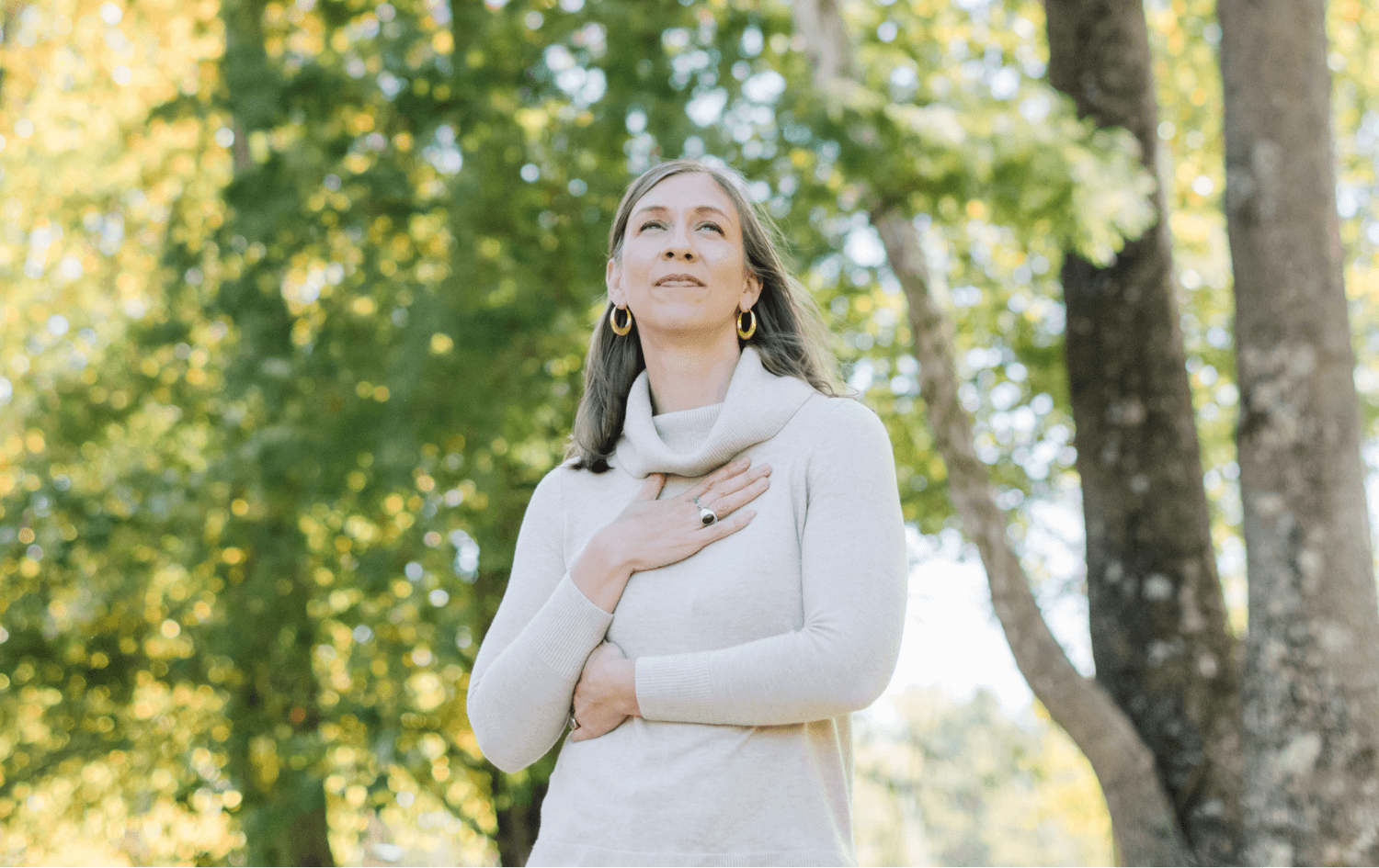 Woman standing outdoors in front of trees with sunlight filtering through, hand on her chest.