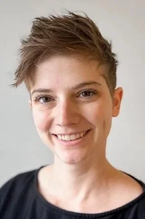 Close-up of a smiling woman with short, tousled brown hair and fair skin, wearing a black top against a plain light background.