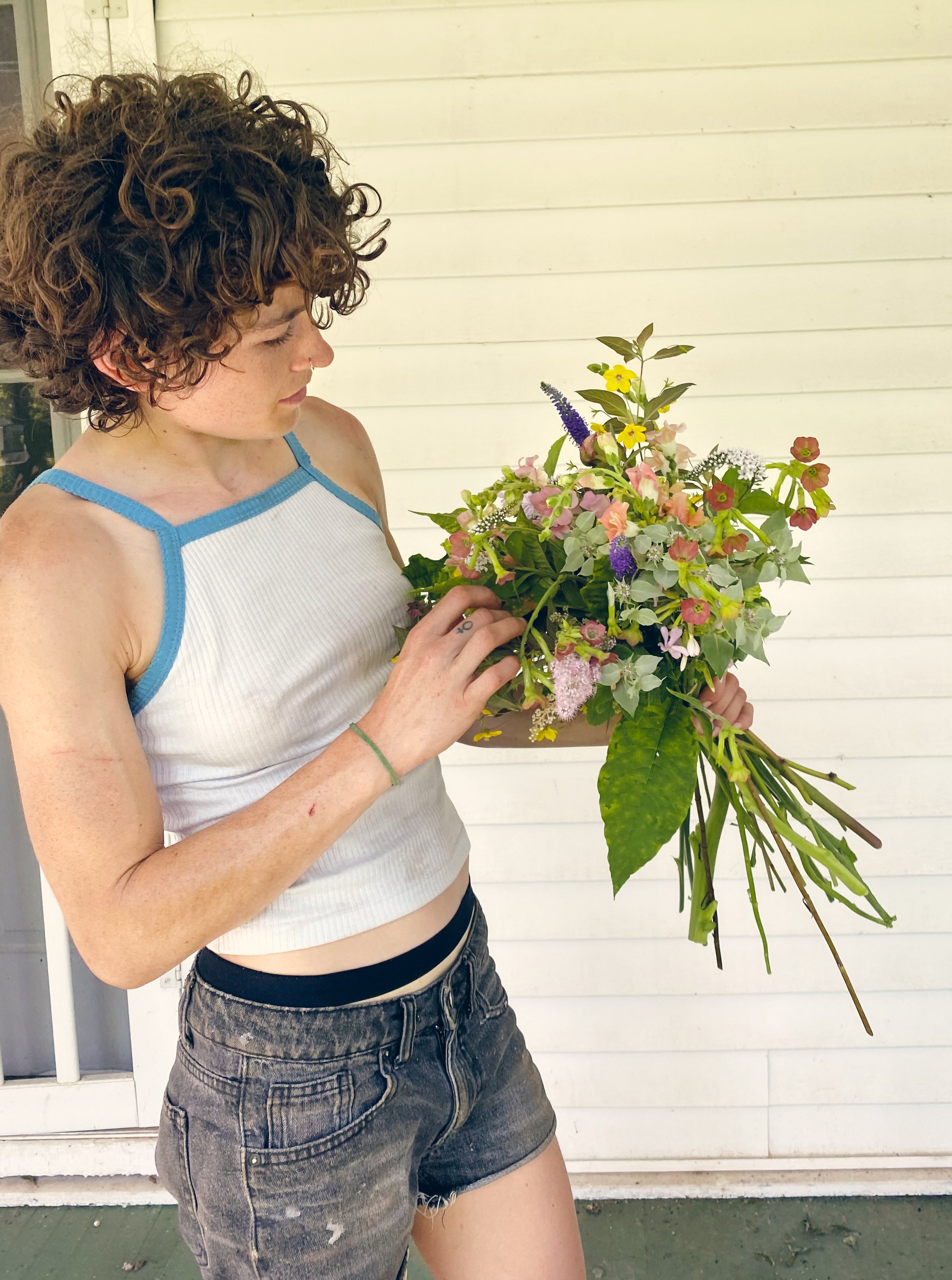 A woman with curly brown hair, wearing a white tank top with blue trim and black shorts, is holding a large bouquet of colorful flowers, standing against a white wooden wall.
