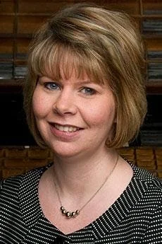 Portrait of a woman with short blonde hair, smiling, wearing a black and white patterned top and a pearl necklace, with a wooden background.