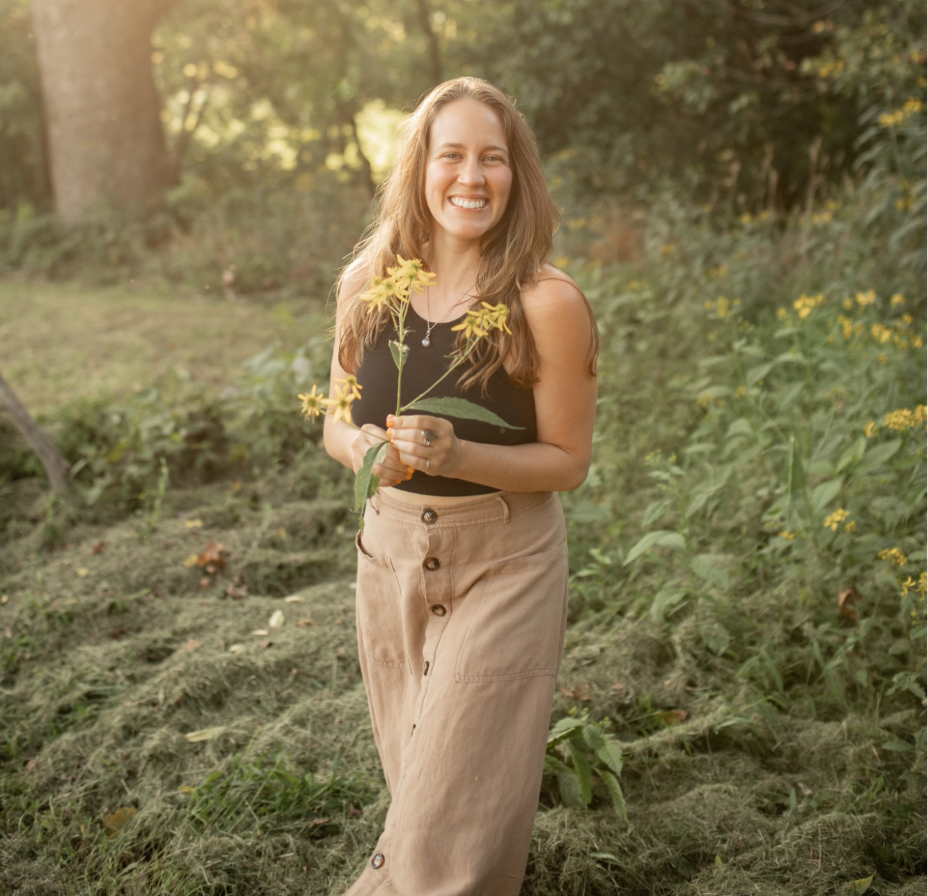 A woman standing outdoors in a natural setting, holding yellow flowers, and smiling at the camera.