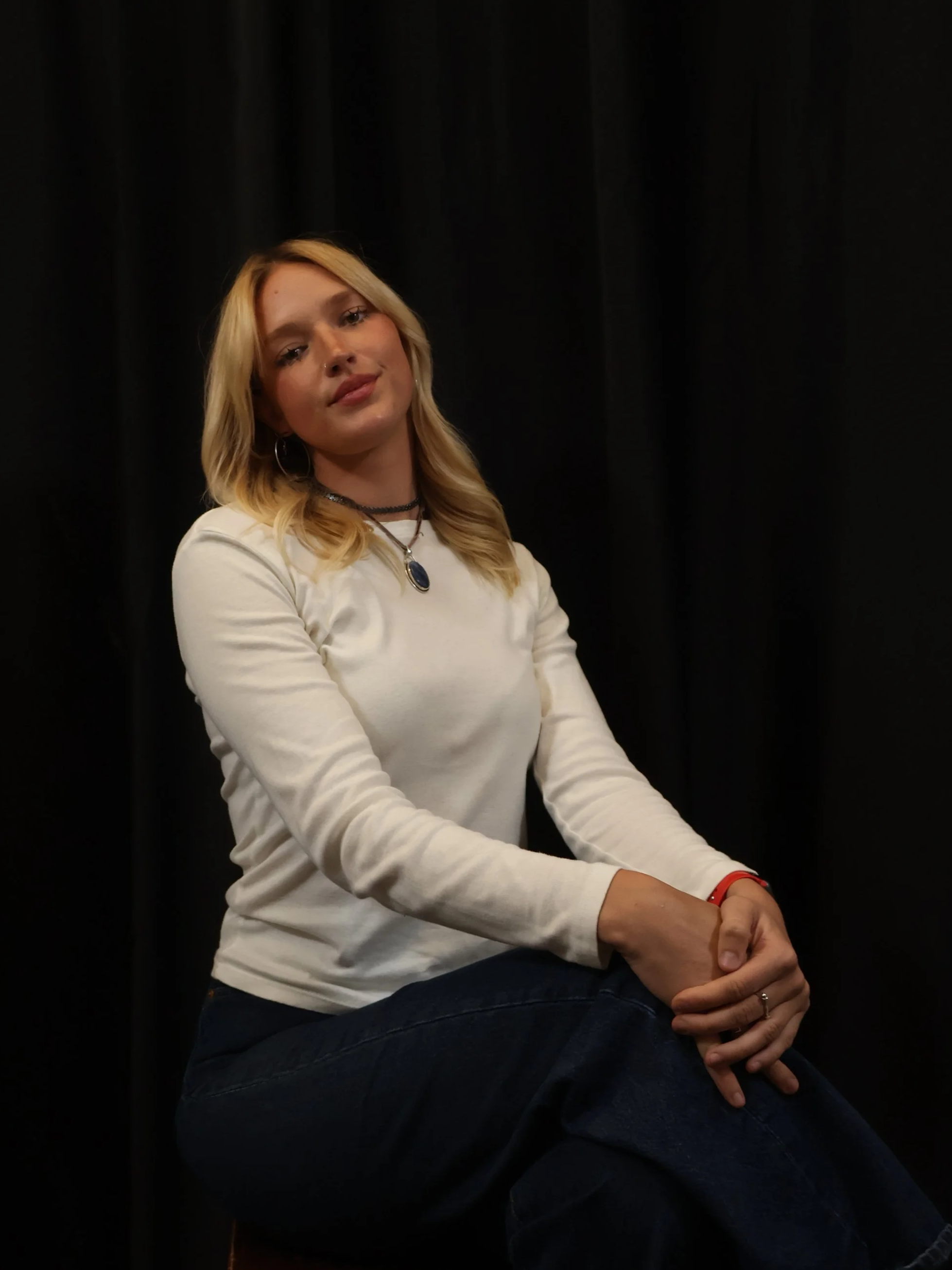 A young woman with blonde hair and hoop earrings, dressed in a white long-sleeve shirt, sitting with crossed arms against a black curtain background.