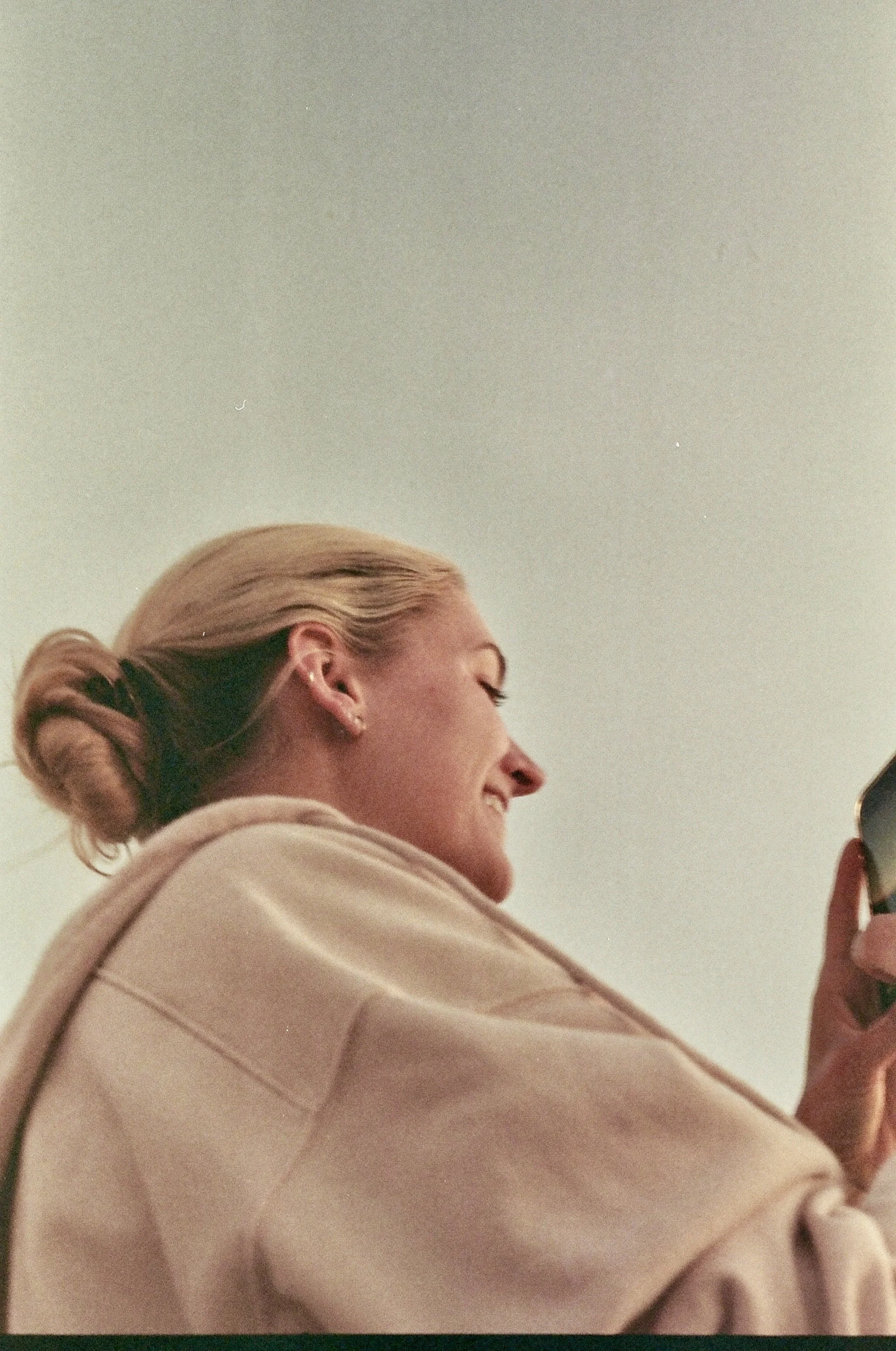 Close-up of a woman with blonde hair styled in a neat bun, wearing a beige hoodie, smiling and looking towards the right.