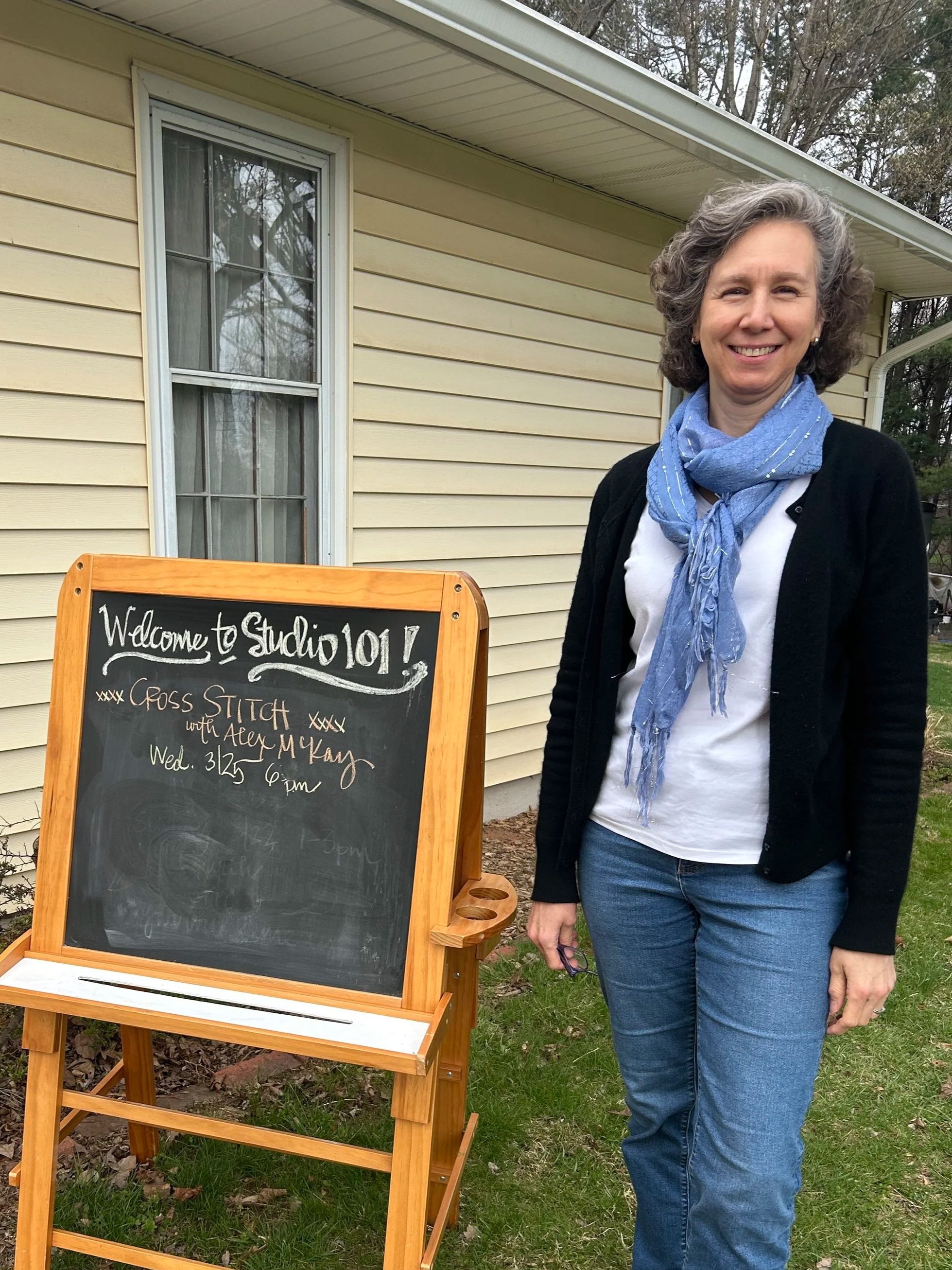 Woman standing outside near a wooden chalkboard sign with a welcome message for Studio 101.7, written with white chalk, in front of a light yellow house with a window, trees in the background, and leafy ground.