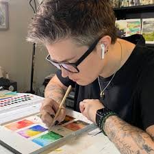 Person with short hair, glasses, and tattoos working on a watercolor painting or color swatch chart, wearing AirPods.